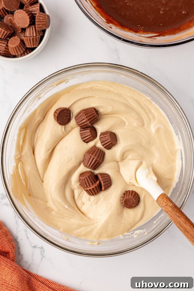 Miniature peanut butter cups are being sprinkled into the peanut butter ice cream mixture in a glass bowl, after the whipped cream has been gently folded in.