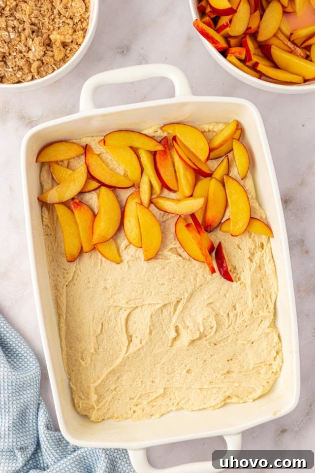 An overhead shot depicting a generous layer of seasoned peaches partially covering the smooth cake batter in a rectangular baking pan, setting the stage for the next delicious layer.