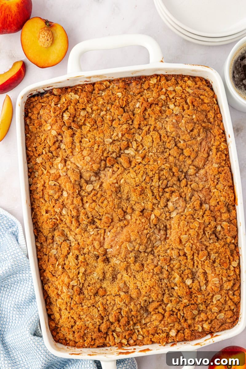 An inviting overhead view of a freshly baked peach coffee cake, golden brown and glistening with a perfect crumb topping, cooling in its rectangular pan after emerging from the oven.