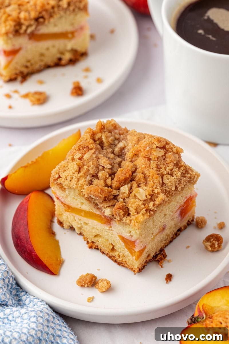 A delightful slice of peach coffee cake resting on a white plate, with another slice and a comforting mug of coffee partially visible in the softly blurred background, suggesting a perfect morning treat.