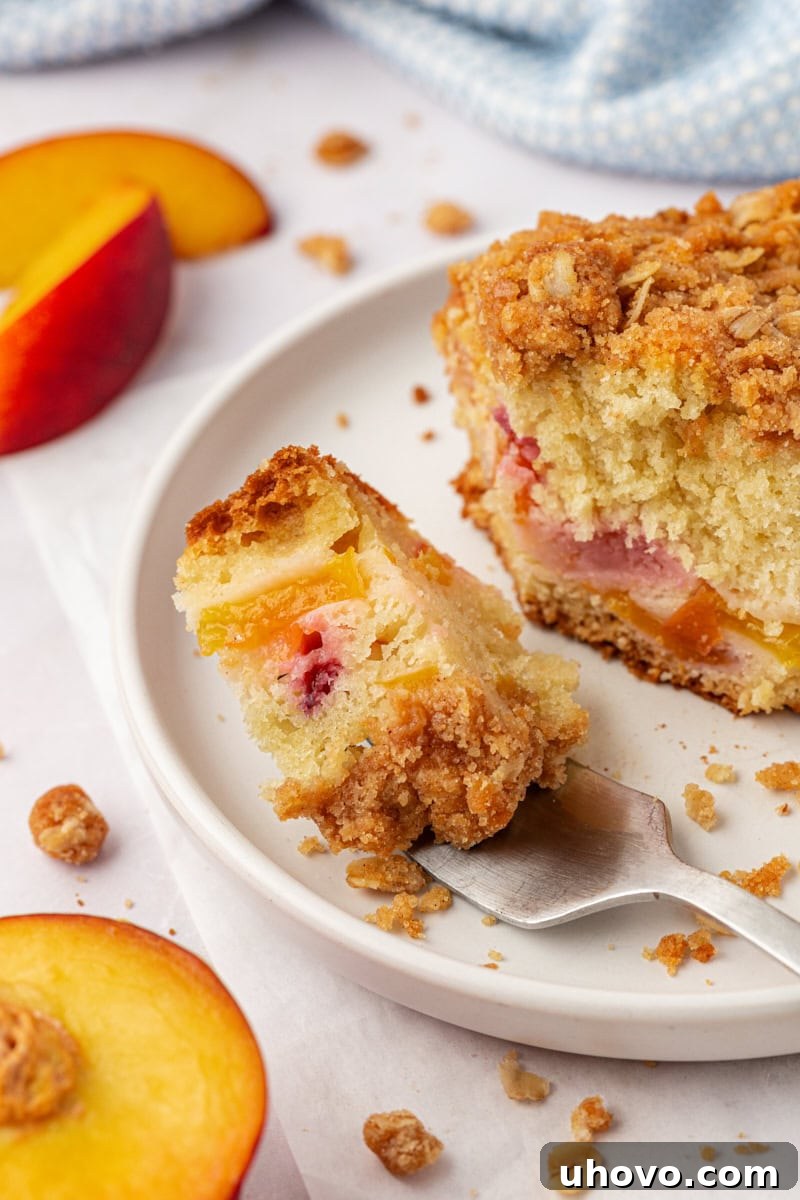 A close-up of a delightful bite of peach coffee cake resting on a fork, poised above a white plate with the remaining cake artfully blurred in the background, showcasing its delicious layers.