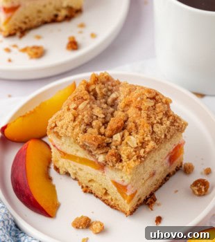 A slice of peach coffee cake on a white plate with another slice of cake and a mug of coffee partially visible in the background