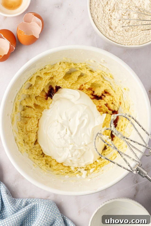 An overhead view showing sour cream and vanilla extract being incorporated into a creamy mixture of beaten butter, sugar, and eggs, creating a smooth and luscious cake batter base.