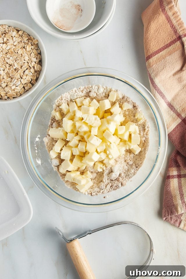 Overhead perspective showing cold, cubed butter being gently added to the dry mixture of flour, brown sugar, cinnamon, and salt in a glass mixing bowl for the crumb topping.