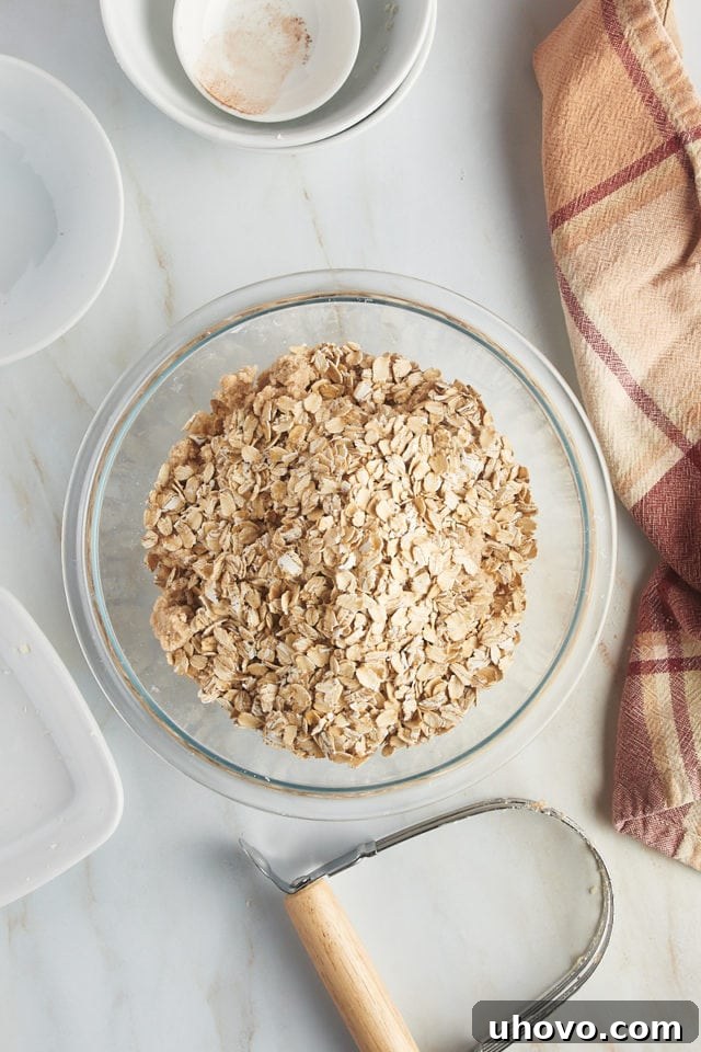 Overhead shot of old-fashioned rolled oats being added to the crumb topping mixture in a glass bowl, signaling the final stage of crumble preparation.