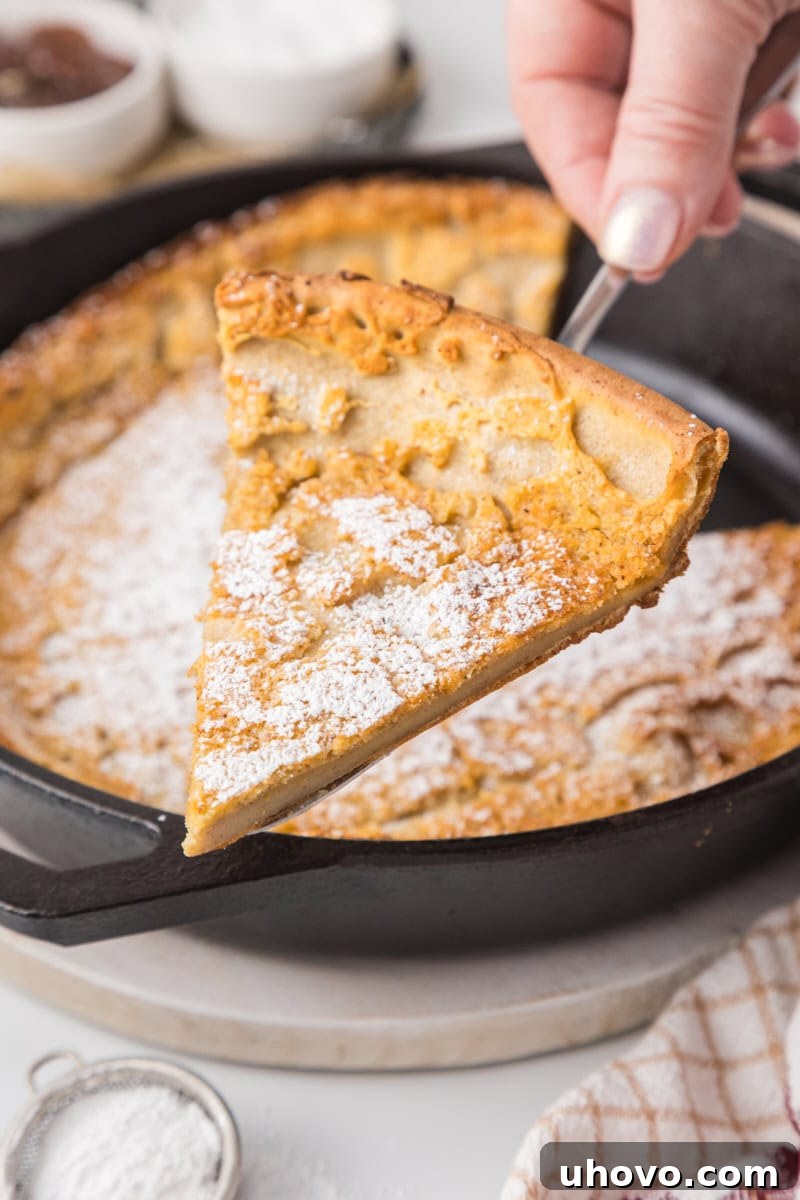 a slice of Dutch baby pancake on a pie server with the remaining puffed pancake still in the cast iron skillet in the background