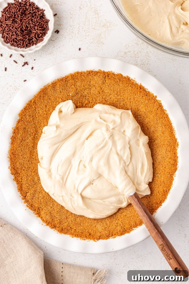 The smooth Bailey's cheesecake filling being evenly spread into the baked graham cracker crust within a white pie plate.