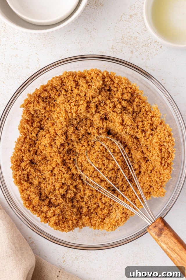 An overhead view of the thoroughly mixed graham cracker crust mixture in a clear glass bowl, showcasing its uniform, moist consistency.