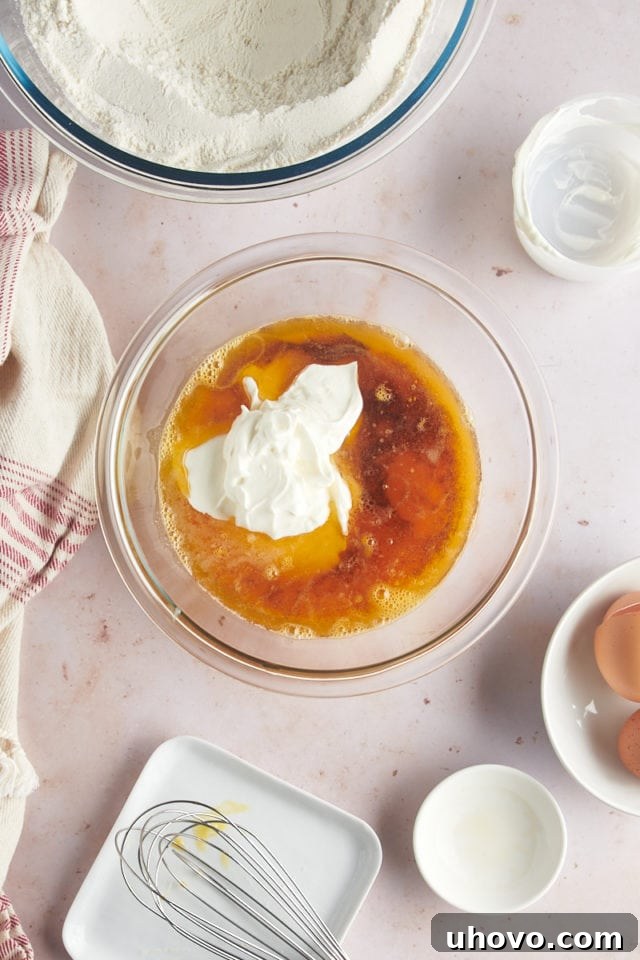 An overhead view of individual wet ingredients—eggs, cooled brown butter, sour cream, and vanilla extract—gathered in a glass mixing bowl before being combined.