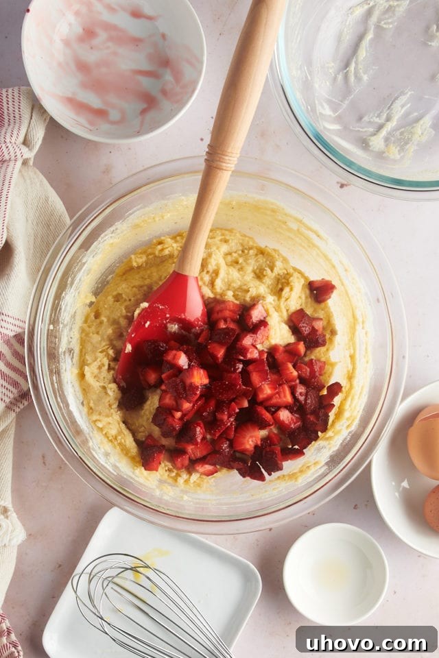 An overhead view of chopped fresh strawberries being gently folded into the muffin batter in a glass mixing bowl, ensuring even distribution without overmixing.