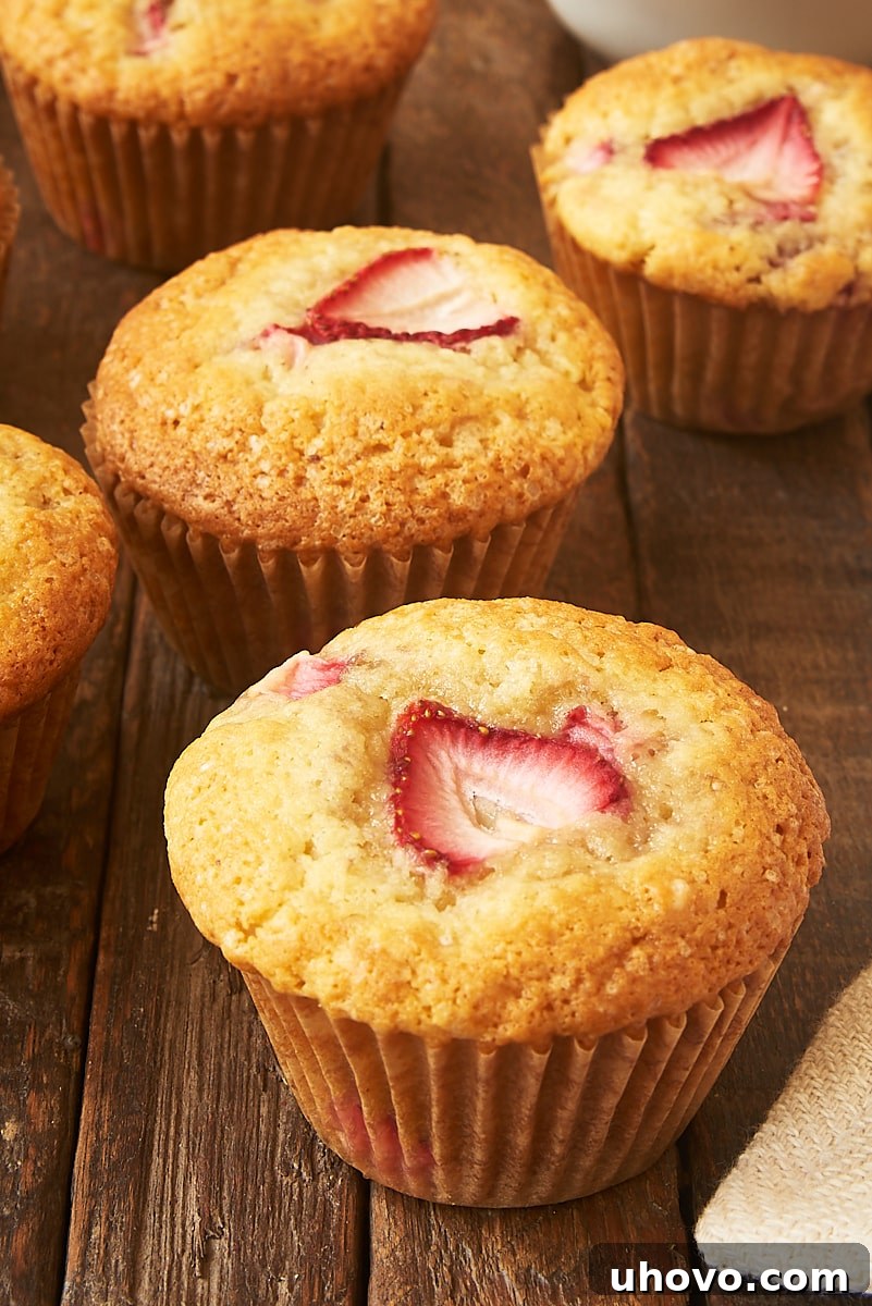 A single, perfectly baked brown butter strawberry muffin placed on a rustic wooden surface, with more muffins gently blurred in the background, highlighting its golden crust and visible strawberry pieces.