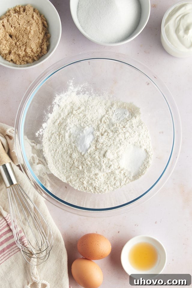 An overhead view of individual dry ingredients—flour, baking powder, baking soda, and salt—placed separately in a glass mixing bowl before being combined.