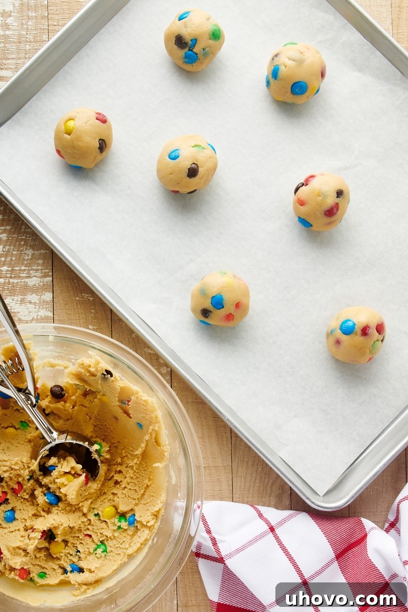 An overhead shot of perfectly uniform M&M cookie dough balls, neatly arranged on a parchment-lined baking sheet, awaiting their bake time.