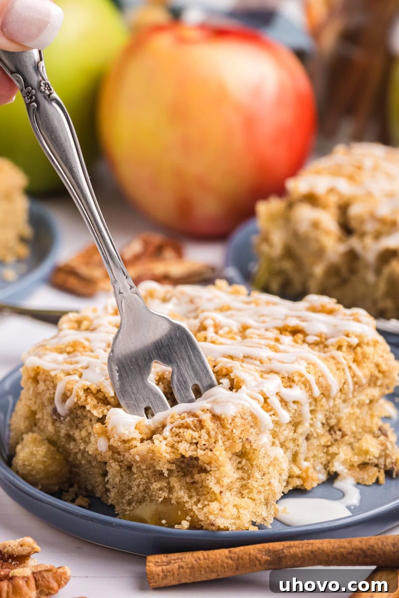 A beautifully presented slice of apple crumb sheet cake resting on a white plate, with a fork gently positioned to take a bite, highlighting the tender interior and the crisp crumb topping.