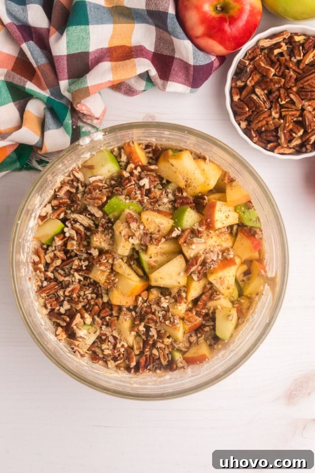 Overhead view of freshly chopped apples and crushed pecans being gently stirred into the rich cake batter in a mixing bowl, ensuring even distribution.