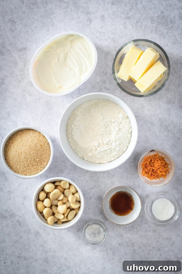 All the essential ingredients for Cream Cheese Macadamia Cookies are neatly laid out on a kitchen counter, including fresh flour, soft butter, cream cheese, brown sugar, fragrant vanilla, a bright orange for zesting, and coarsely chopped macadamia nuts.