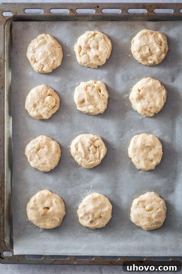 An overhead shot revealing perfectly scooped portions of Cream Cheese Macadamia Cookie dough, dotted with nuts, neatly arranged on a parchment-lined baking sheet, patiently awaiting their turn in the oven.