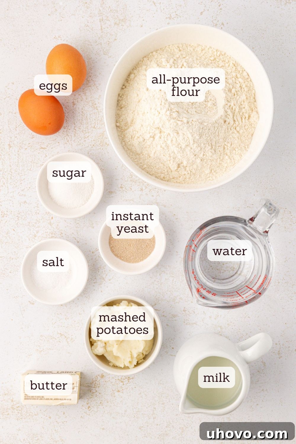 An overhead shot showcasing all the essential ingredients laid out for making potato rolls: flour, yeast, sugar, salt, milk, water, butter, mashed potatoes, and eggs.