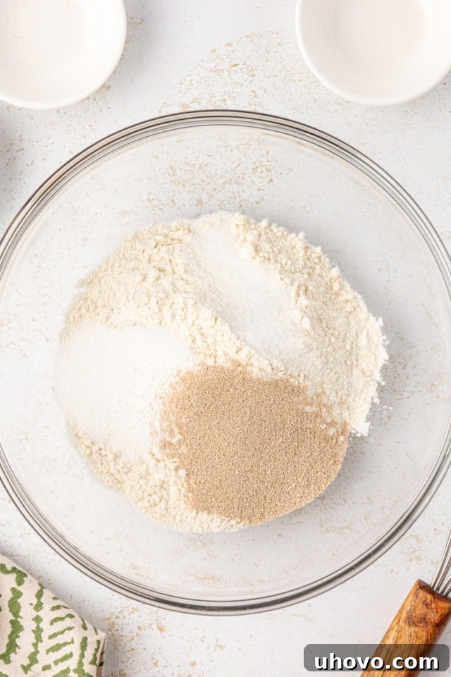 An overhead view of a glass mixing bowl containing all-purpose flour, sugar, instant yeast, and salt, perfectly portioned for the first step of the potato roll recipe.