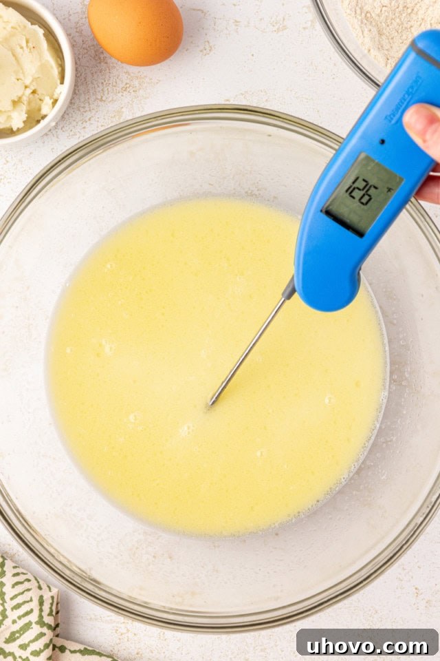 A close-up, overhead view of a saucepan with a mixture of heated water, milk, and melted butter. An instant-read thermometer indicates the liquid has reached 126°F, the ideal temperature for yeast activation.
