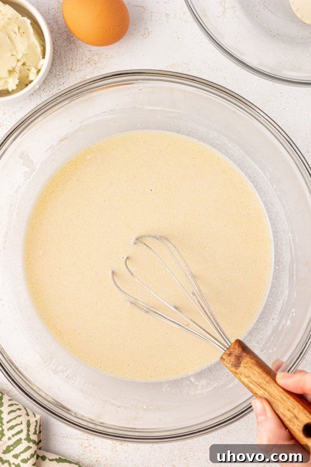 An overhead shot of a glass mixing bowl showing the initial stages of dough formation for potato rolls, where the warm liquid mixture has just been stirred into the dry flour and yeast mixture.