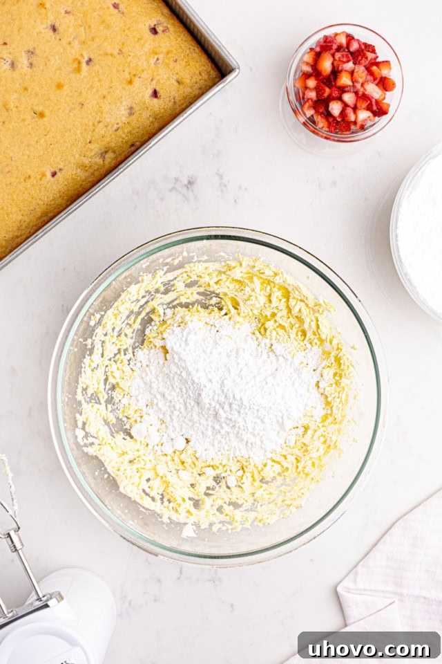 An overhead view of confectioners' sugar being carefully sifted and added to a creamy mixture of beaten butter and cream cheese in a glass mixing bowl, a key stage in preparing the frosting.