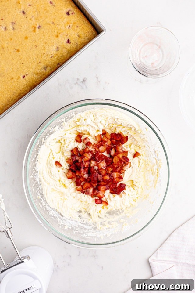 A close-up overhead shot depicting vibrant chopped strawberries being gently stirred into a creamy, pale pink cream cheese frosting, highlighting the final flavor infusion for the strawberry sheet cake.
