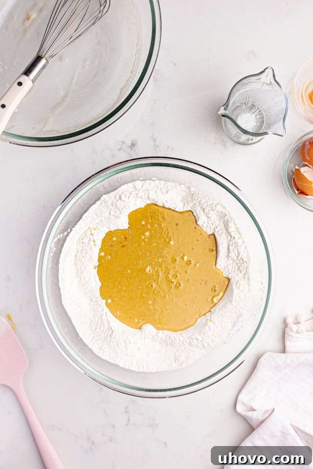 A close-up overhead view illustrating the wet ingredients being carefully poured into the well of the dry ingredients for the strawberry sheet cake, signaling the crucial step of combining the mixtures to form the batter.
