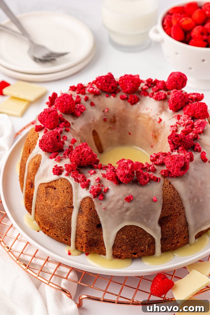 A beautifully decorated White Chocolate Raspberry Bundt Cake on a white cake plate, with fresh raspberries and additional plates subtly visible in the soft-focus background.
