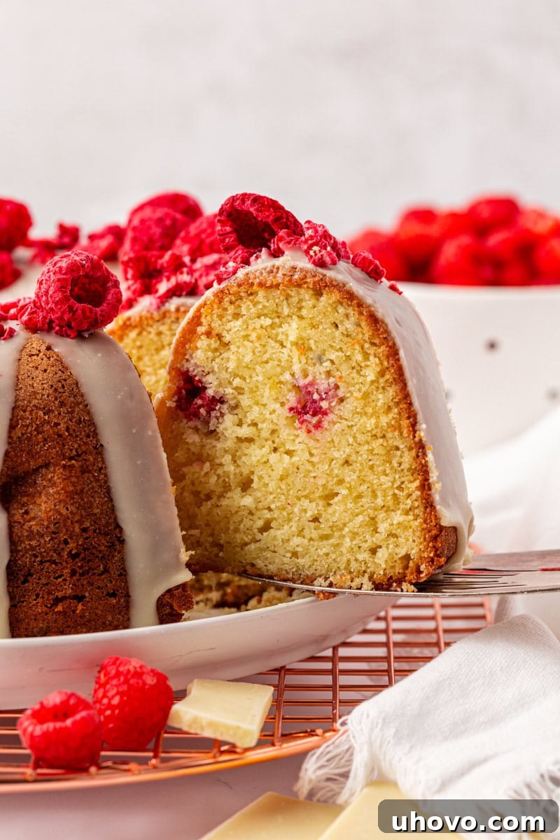 A slice of White Chocolate Raspberry Bundt Cake being lifted away from the main cake on a cake server, revealing its moist, berry-filled interior.