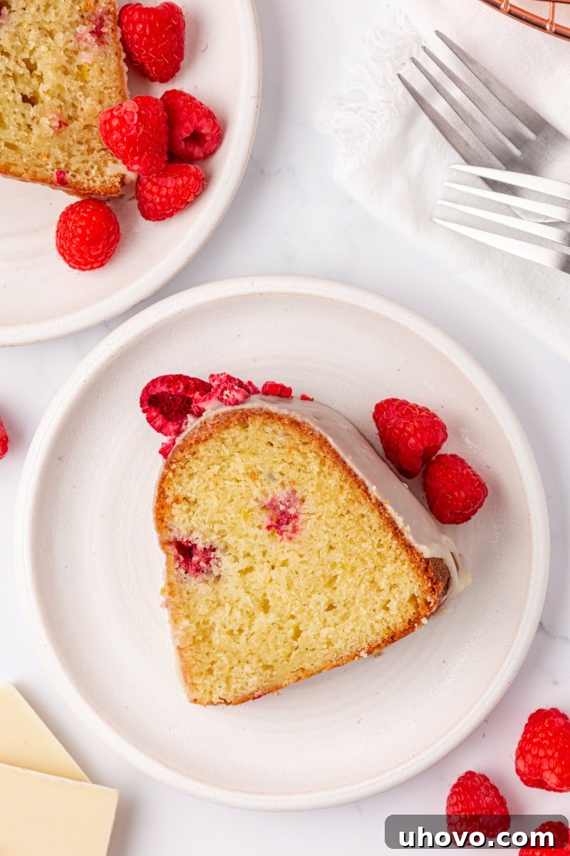 An overhead view of two slices of White Chocolate Raspberry Bundt Cake on white plates, accompanied by fresh raspberries, forks, and scattered white chocolate pieces, creating an inviting dessert scene.