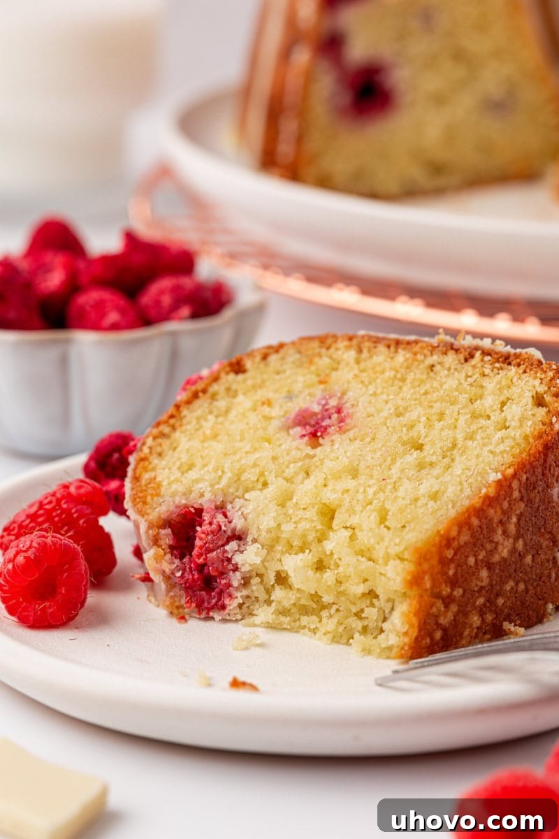 A close-up of a slice of White Chocolate Raspberry Bundt Cake with a bite taken out, showing the moist interior and raspberries. The rest of the cake and a bowl of fresh raspberries are blurred in the background.