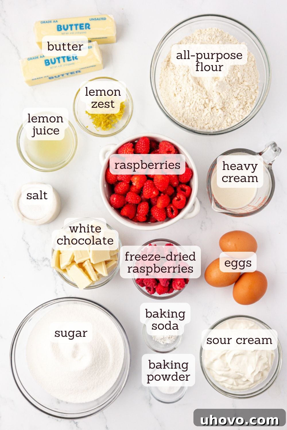An overhead shot of various ingredients for White Chocolate Raspberry Bundt Cake laid out on a clean surface, including flour, sugar, butter, eggs, raspberries, and a lemon.