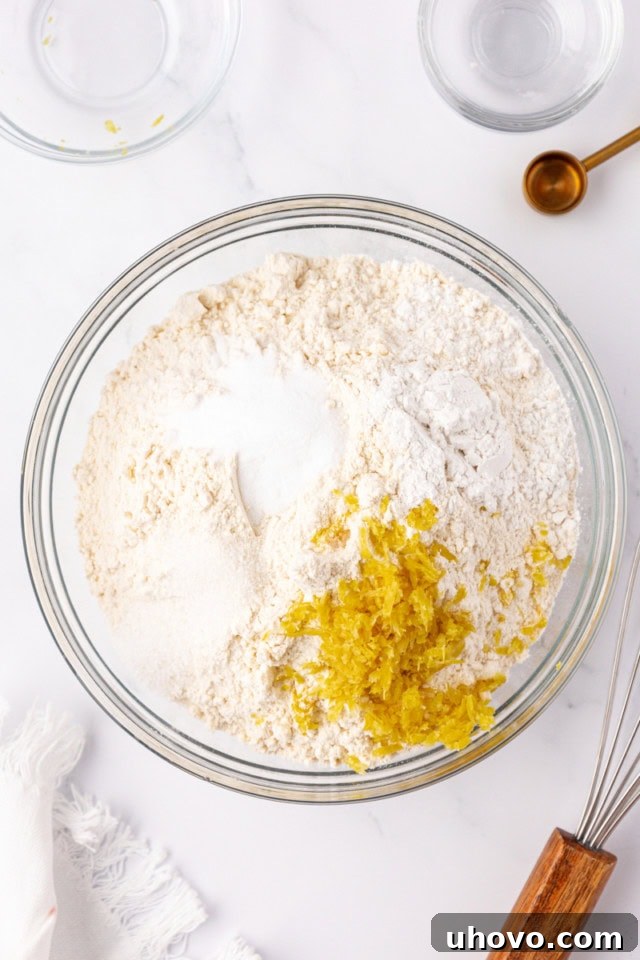 An overhead view of flour, baking powder, baking soda, salt, and fresh lemon zest perfectly combined in a clear glass mixing bowl, representing the dry ingredients step.