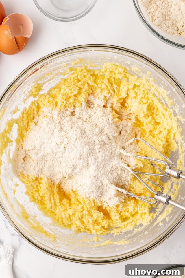 Dry flour mixture being gradually added to the wet cake batter in a glass mixing bowl, showing the alternate addition technique.