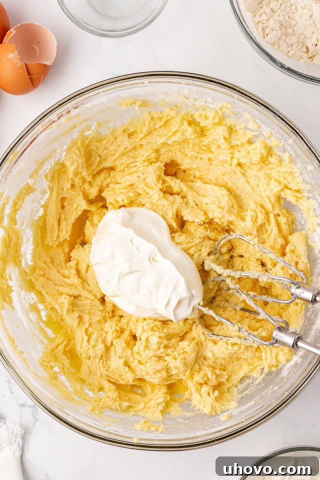 A dollop of sour cream being added to the cake batter in a glass mixing bowl, illustrating the alternating addition with dry ingredients.