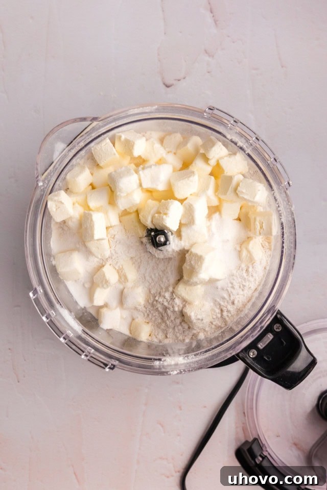 overhead view of butter, cream cheese, flour, sugar, and salt in the bowl of a food processor