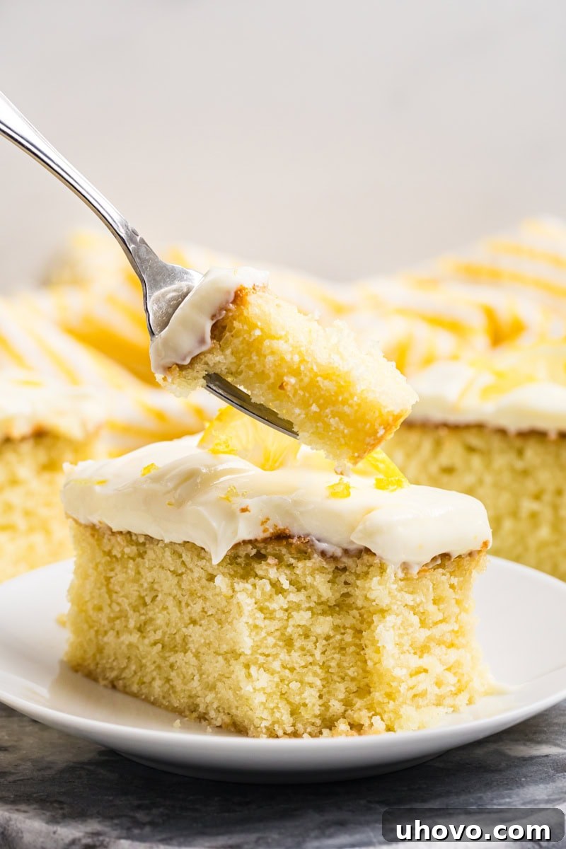 A delicious bite of lemon sheet cake on a fork, poised above the remaining slice of cake on a plate.