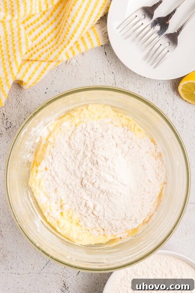 Overhead view of dry ingredients being gradually added to the lemon cake batter in a mixer bowl.
