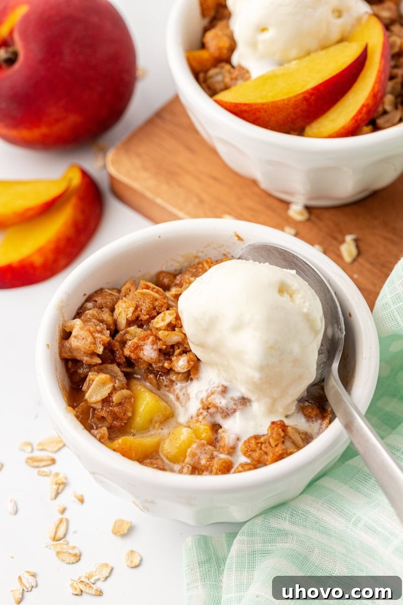 A warm peach crisp topped with a scoop of vanilla ice cream, served in a white bowl with fresh peaches and another serving blurred in the background, evoking a comforting summer dessert scene.