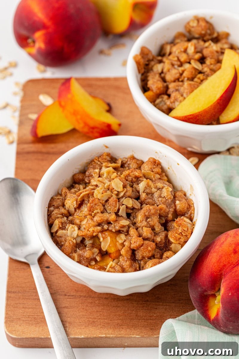 Two servings of peach crisp, one on a wooden board with a spoon and the other in a bowl, surrounded by fresh peaches, creating a rustic and inviting display.