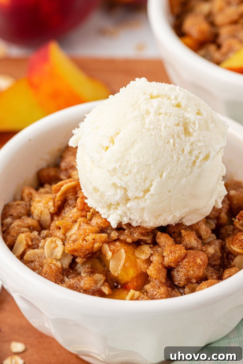 A close-up view of a serving of peach crisp in a white bowl, generously topped with melting vanilla ice cream, highlighting the warm, gooey fruit and crunchy topping.