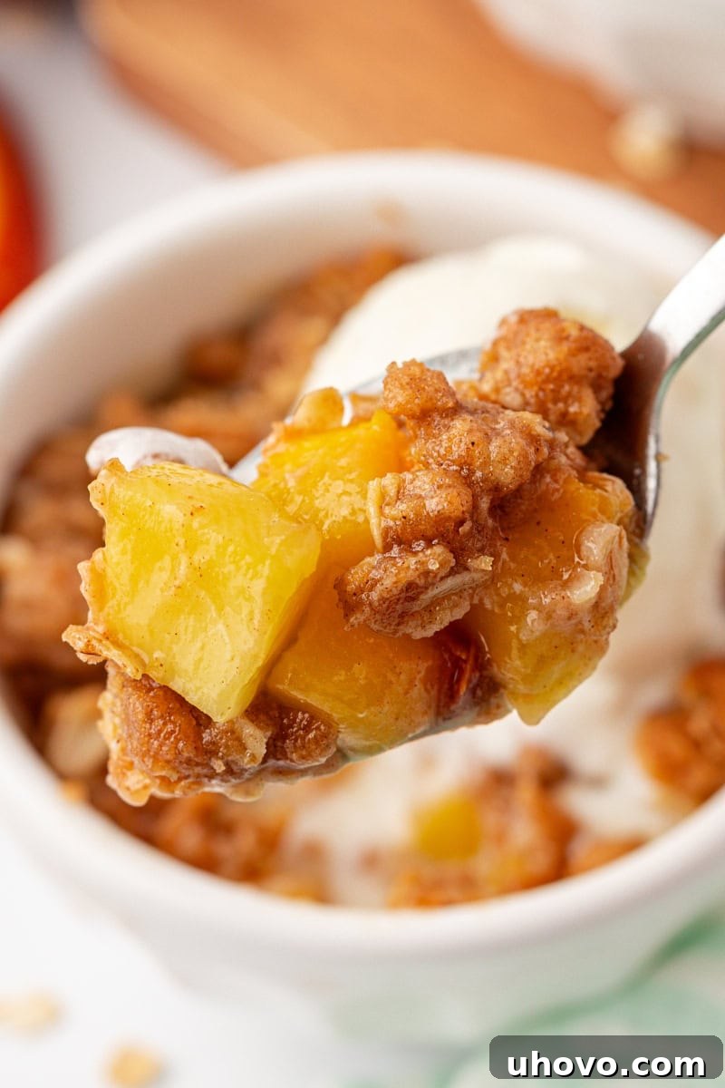 A close-up shot of a spoon holding a perfect bite of peach crisp, showing the warm, syrupy fruit and crumbly topping, with the remaining dessert blurred in the background.