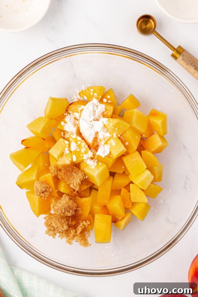 An overhead view of chopped peaches, brown sugar, cornstarch, and almond extract being mixed in a clear glass bowl, illustrating the first step of preparing the crisp filling.