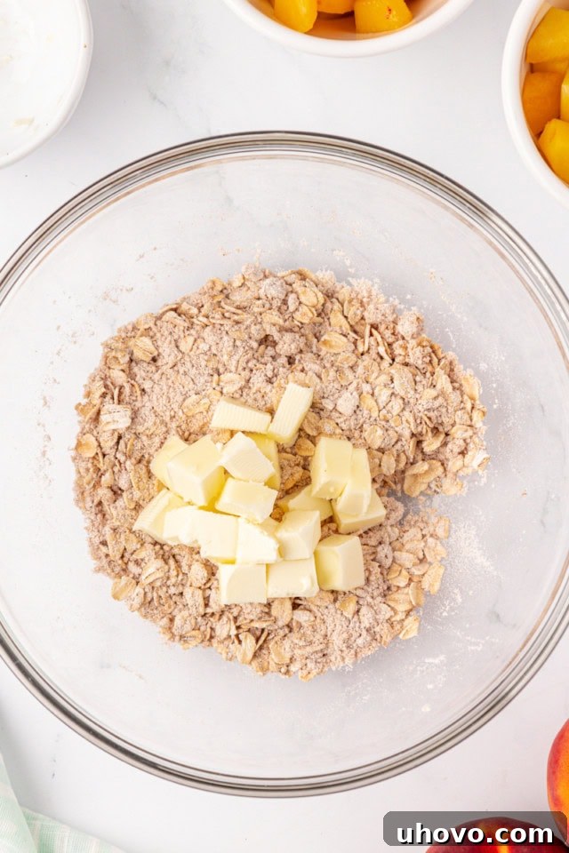 An overhead shot of cold butter cubes being added to a mixture of oats, flour, and brown sugar in a glass bowl, illustrating the initial step of preparing the crisp topping.