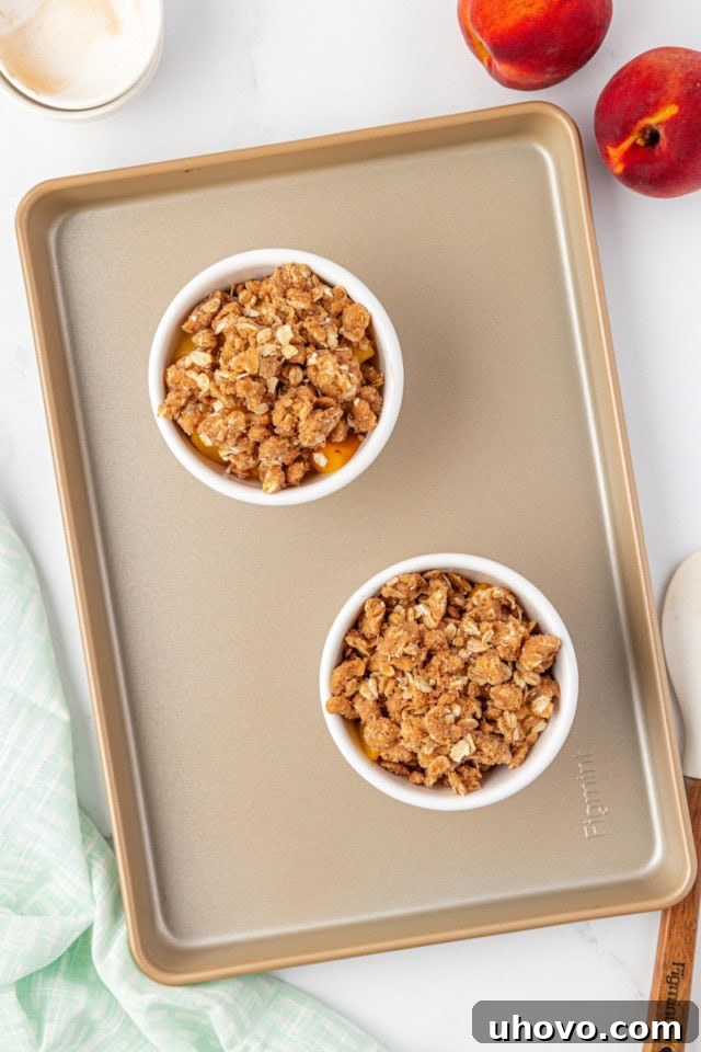 An overhead view of two small baking dishes filled with peach crisp filling, generously topped with the crumbly crisp mixture, placed on a baking sheet, ready for the oven.