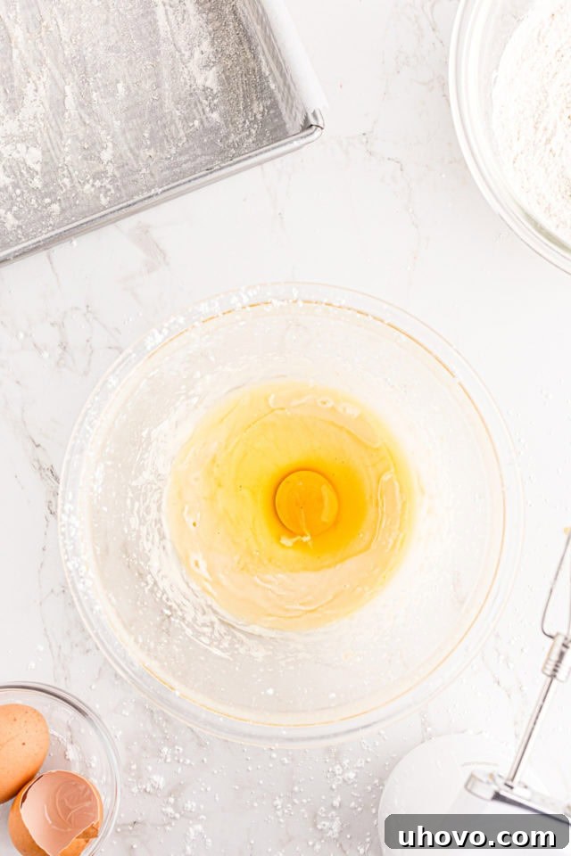 Overhead view of an egg being added to the creamy batter mixture in a glass bowl, illustrating the process of mixing the blackberry crumb bar base.