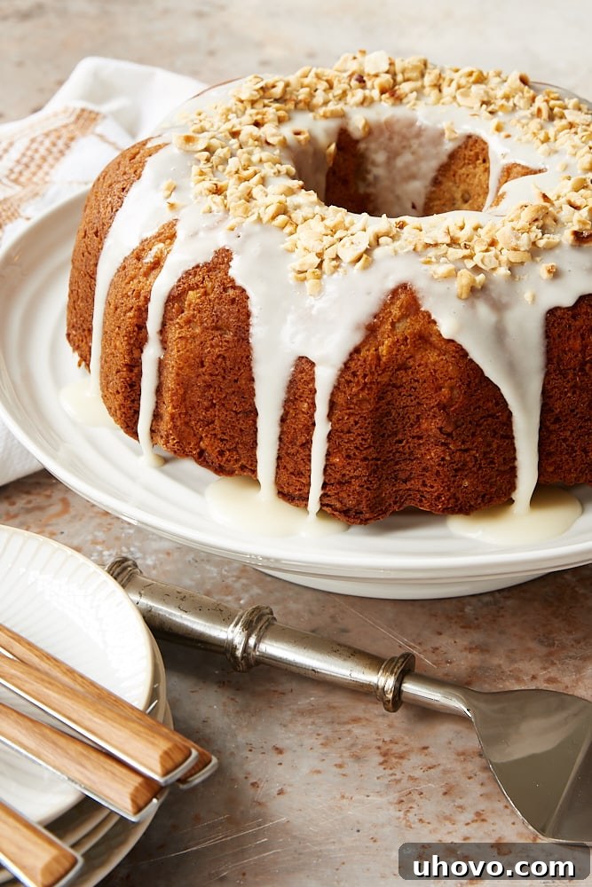 A beautifully baked Hazelnut Pear Bundt Cake on a white cake stand, showcasing its elegant fluted design and a delicate sour cream glaze drizzling down the sides. It's garnished with a few chopped hazelnuts.