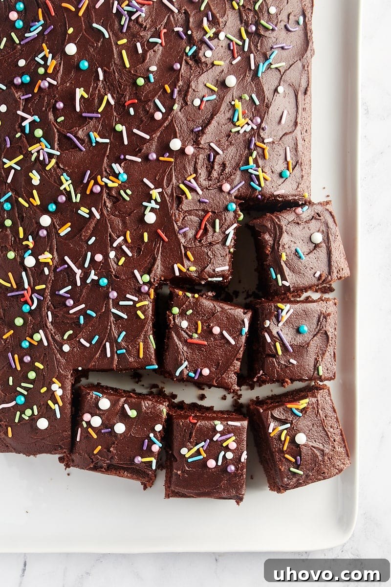 An overhead view of a partially sliced, wonderfully rich and moist Chocolate Sheet Cake on a pristine white serving tray, showcasing its smooth frosting and inviting texture.