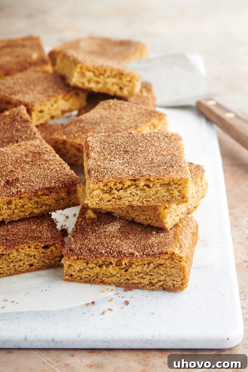 A mouth-watering stack of golden Snickerdoodle Bars, dusted with cinnamon sugar, arranged on a rustic white board, inviting a taste.